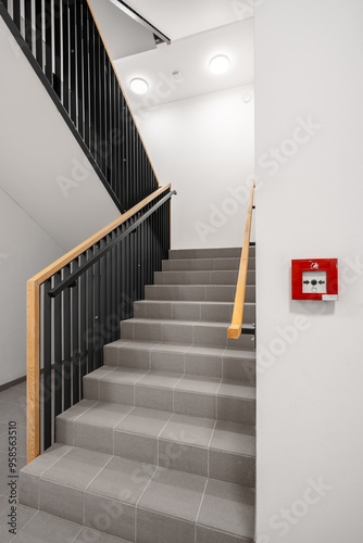 A modern staircase with black metal railings, wooden handrails, and tiled steps, located inside a building. The walls are white, and there is an emergency fire alarm on the right.