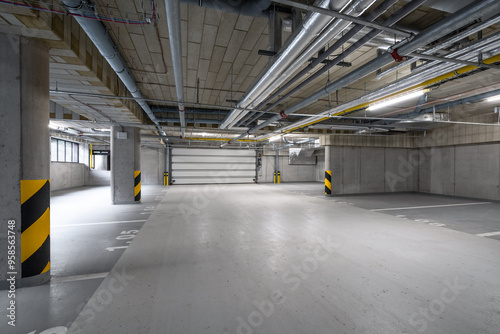 A spacious underground parking garage with exposed pipes on the ceiling, yellow and black striped pillars, concrete walls, and marked parking spaces, with no cars or people present.