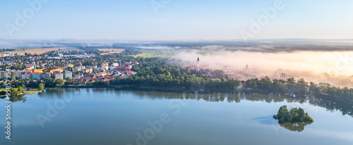 Aerial perspective captures Trebon enveloped in morning haze. The tranquil waters reflect the fog, highlighting the blend of historic architecture and lush landscapes, inviting a sense of peace and