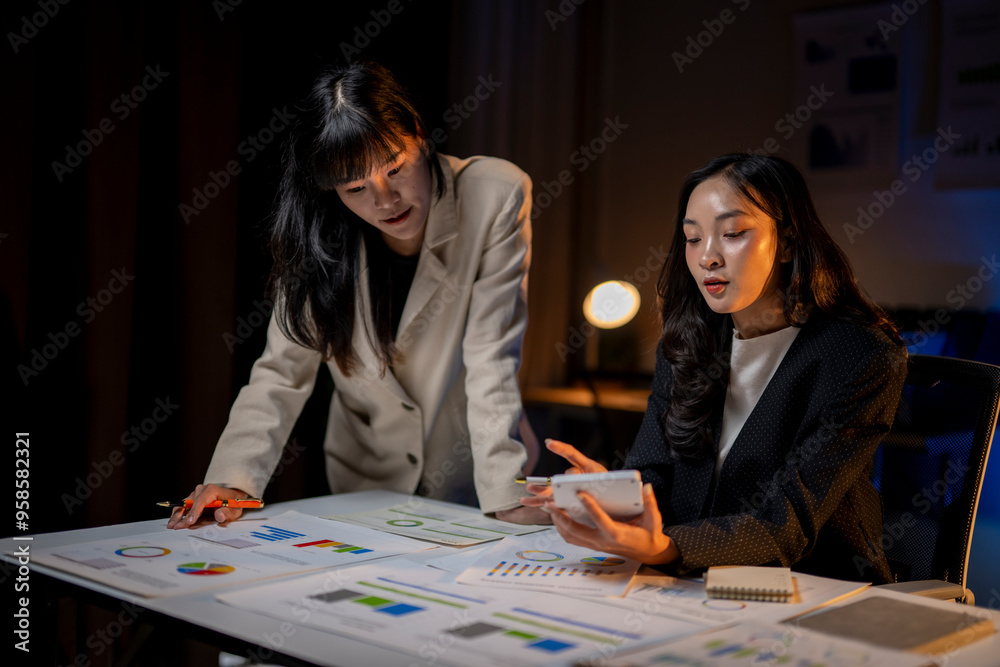 Fototapeta premium Two businesswomen working late analyzing financial charts and using calculator