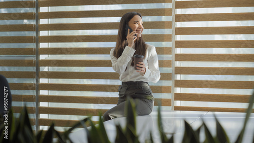 Happy smiling laughing Caucasian woman girl business lady businesswoman talking mobile phone call friendly conversation sitting on table talk smartphone drinking coffee looking at window in office