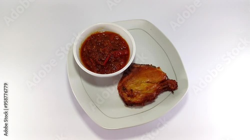 A single piece of fried chicken served alongside red Indonesian chili sambal in a white glass bowl, arranged on a square white plate against a white background, creating a vibrant and appetizing dish photo