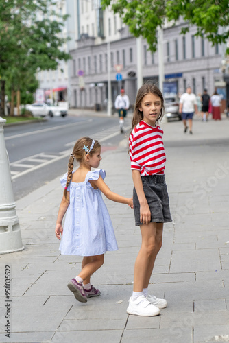 Two young girls stroll along a sidewalk, with one playfully tugging at the other's shorts on a sunny day in the city