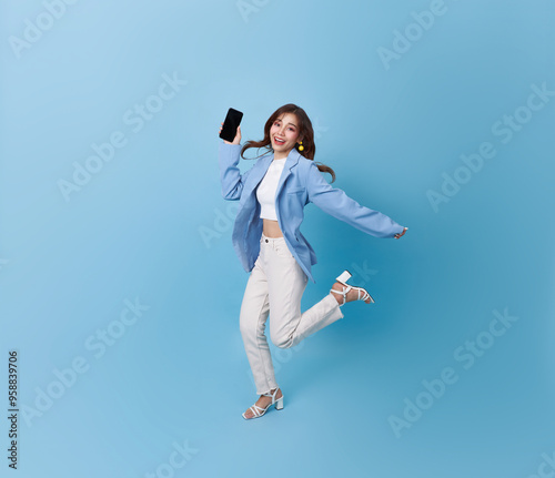 Happy young beautiful Asian business woman showing smartphone blank screen mockup and running jumping isolated on blue background.