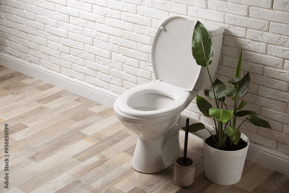 Ceramic toilet bowl, brush and houseplant near white brick wall in restroom. Space for text Ceramic toilet bowl, brush and houseplant near white brick wall in restroom. Space for text