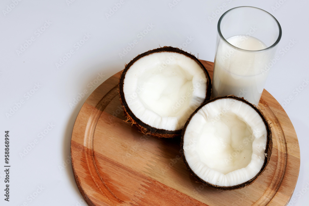 Coconut halves with pulp next to a glass of coconut milk on top of a round wooden board Coconut halves with pulp next to a glass of coconut milk on top of a round wooden board