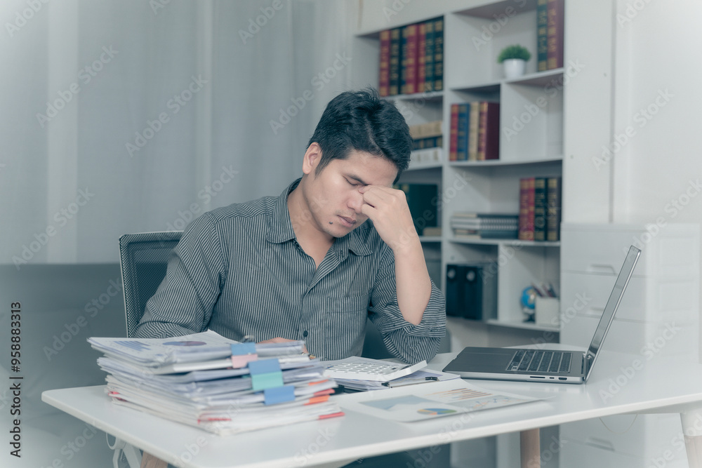 Young Asian man suffering from pain from working at desk, feeling tired from working on laptop and financial documents in office for a long time, Conceptual Office Syndrome