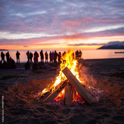 A bonfire burns brightly on a beach at sunset, with a group of people silhouetted in the background.