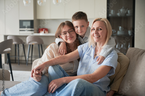 Lovely family relaxing on couch in living room. Little boy hugging his sister and mom. Cozy spending time with family indoors. In background, modern kitchen, neutral colors, minimalistic design.