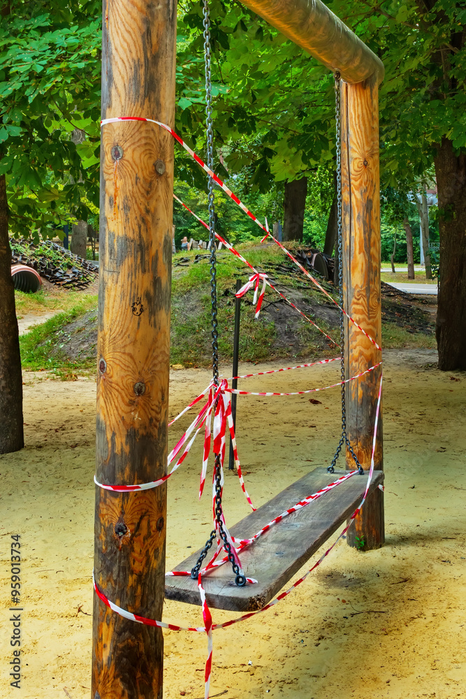Fototapeta premium Wooden swing on chains on empty playground in park. White and red warning tape. Prohibited to use