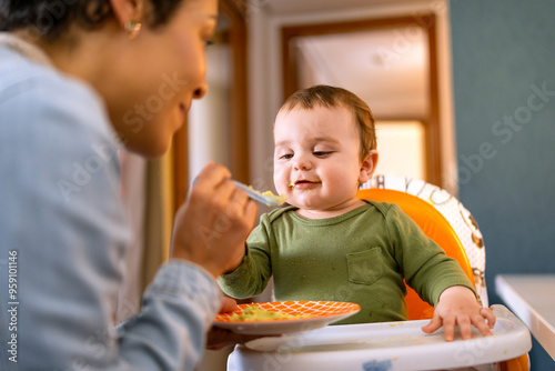 Joyful baby boy in green outfit being spoon-fed by loving mother. Adorable toddler enjoying a healthy meal in high chair at home..