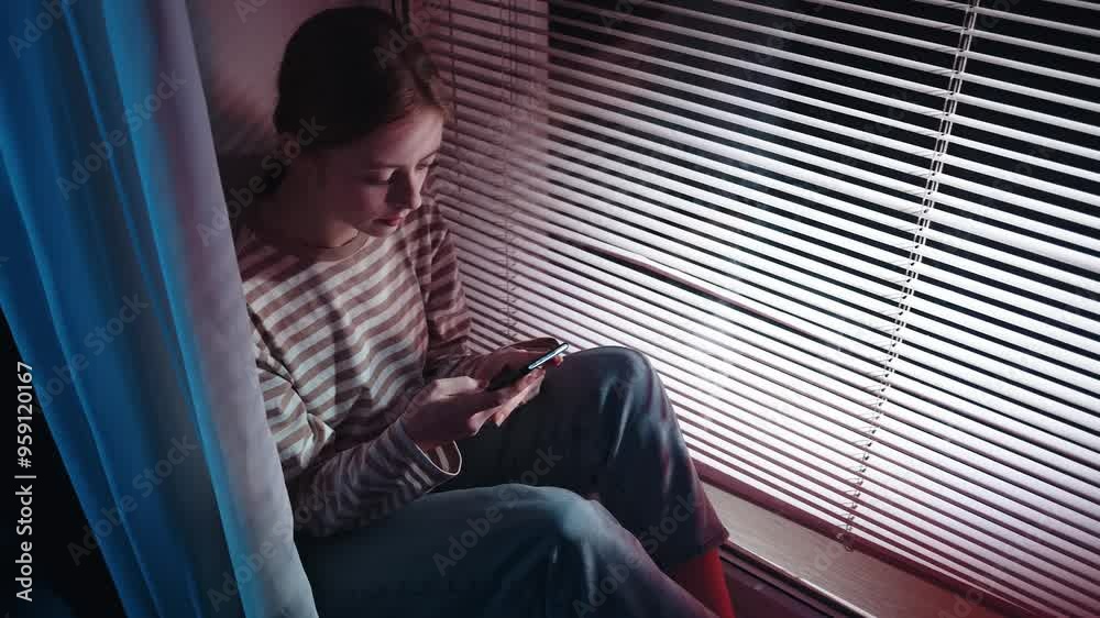 A young, attractive red-haired woman sits by the window, typing on her phone with a light smile and positive mood, enjoying a cozy evening at home A young, attractive red-haired woman sits by the window, typing on her phone with a light smile and positive mood, enjoying a cozy evening at home