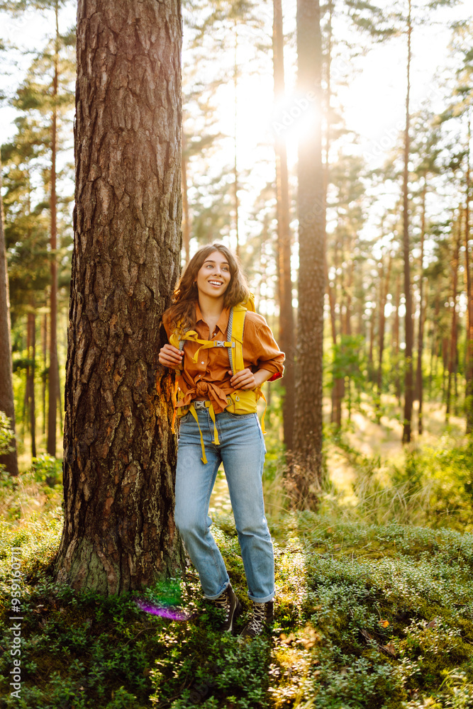 A young woman with curly hair enjoys a serene hike in a sunny forest during the late afternoon, carrying a yellow backpack on her shoulders. The concept of tourism, relaxation, youth and nature. A young woman with curly hair enjoys a serene hike in a sunny forest during the late afternoon, carrying a yellow backpack on her shoulders. The concept of tourism, relaxation, youth and nature.