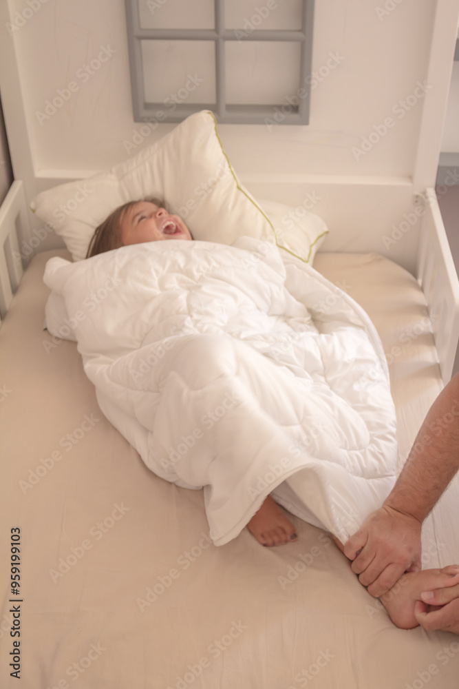 Little girl wrapped in a white blanket while sitting on the bed and smiling being tickled by dad Little girl wrapped in a white blanket while sitting on the bed and smiling being tickled by dad