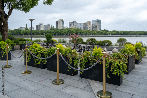 A modern outdoor seating area with rows of planters filled with ornamental plants, separated by golden stanchions with velvet ropes. The space offers a view of the city skyline over the river. photo