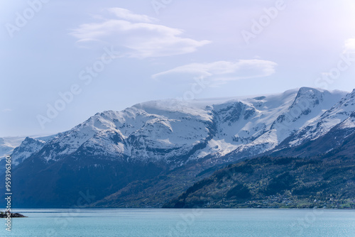 A serene view of the Hardangerfjord. The turquoise waters reflect the surrounding snow-capped mountains and lush green forests. A peaceful scene perfect for relaxation and escape. Voss, Norway.