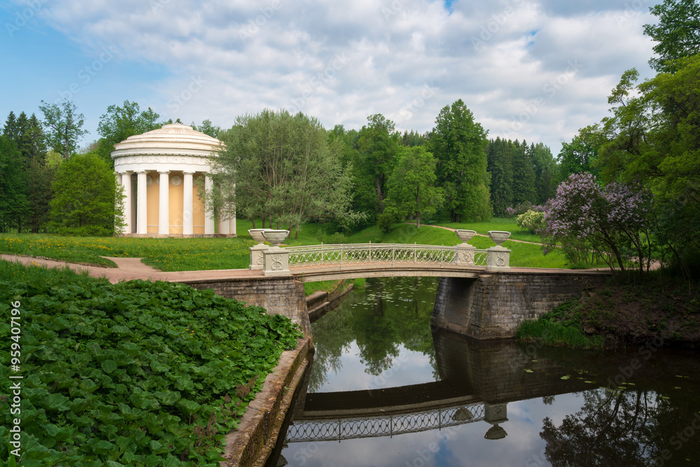Fototapeta premium View of the Temple of Friendship on the banks of the Slavyanka River against the background of the Cast-iron bridge in Pavlovsky Park on a sunny summer day, Pavlovsk, St. Petersburg, Russia