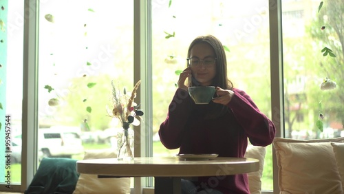 A young woman in glasses talking on the phone and drinking coffee in a cafe with a panoramic window. Free Download A young woman in glasses talking on the phone and drinking coffee in a cafe with a panoramic window. - GFXLoad