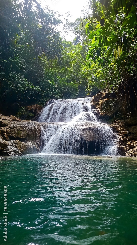Fototapeta premium A cascading waterfall flows over rocks into a still, teal pool in a lush green jungle.