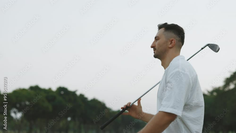 Serious male athlete walking with golf club along course at sunset twilight portrait. Professional player holds metal driver in park against cloudy sky. Sportsman in white shirt at training Serious male athlete walking with golf club along course at sunset twilight portrait. Professional player holds metal driver in park against cloudy sky. Sportsman in white shirt at training
