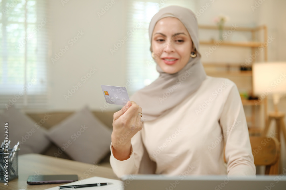 A woman wearing a hijab is holding a credit card in front of a laptop A woman wearing a hijab is holding a credit card in front of a laptop