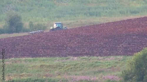 A tractor plows in a field, a rural landscape.