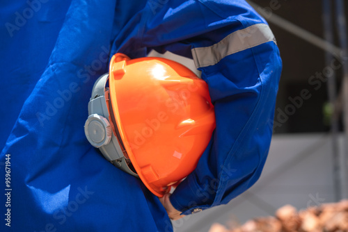 Action of a house builder worker in blue coverall uniform and holding an orange safety hardhat is standing in front of a building house. Ready to working in construction industrial concept scene.