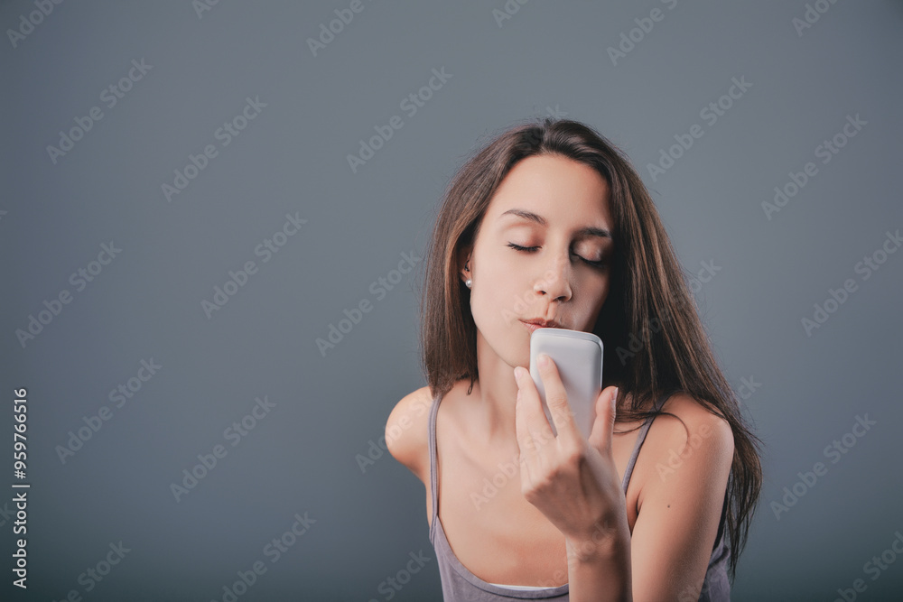 Young woman kissing smartphone with eyes closed on grey backgrou Young woman kissing smartphone with eyes closed on grey backgrou