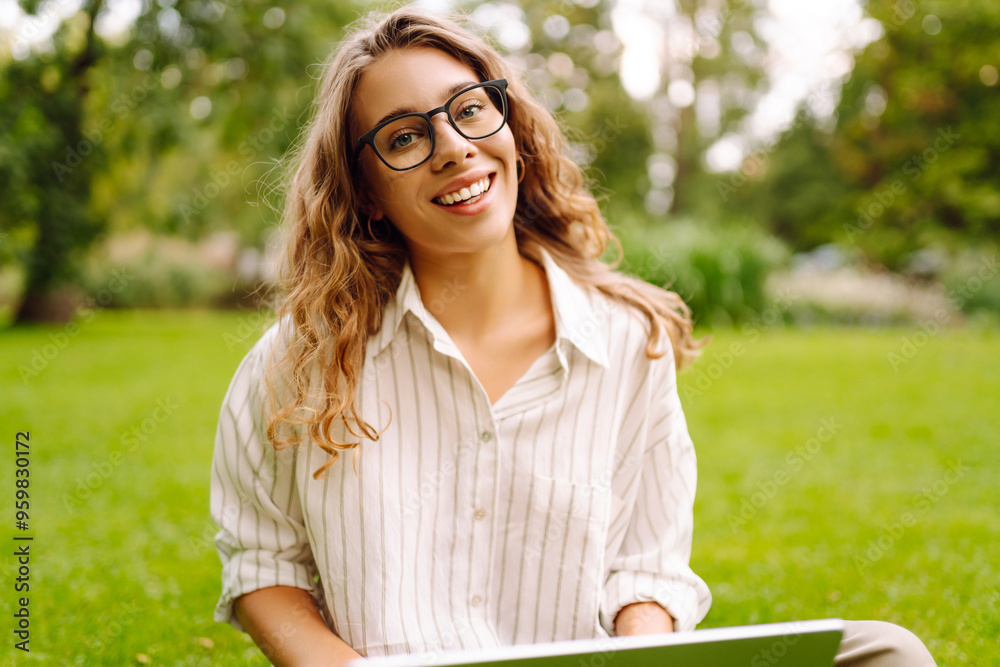 A young woman with curly hair sits comfortably on the grass, using her laptop outdoors. Business, freelancer, online education.