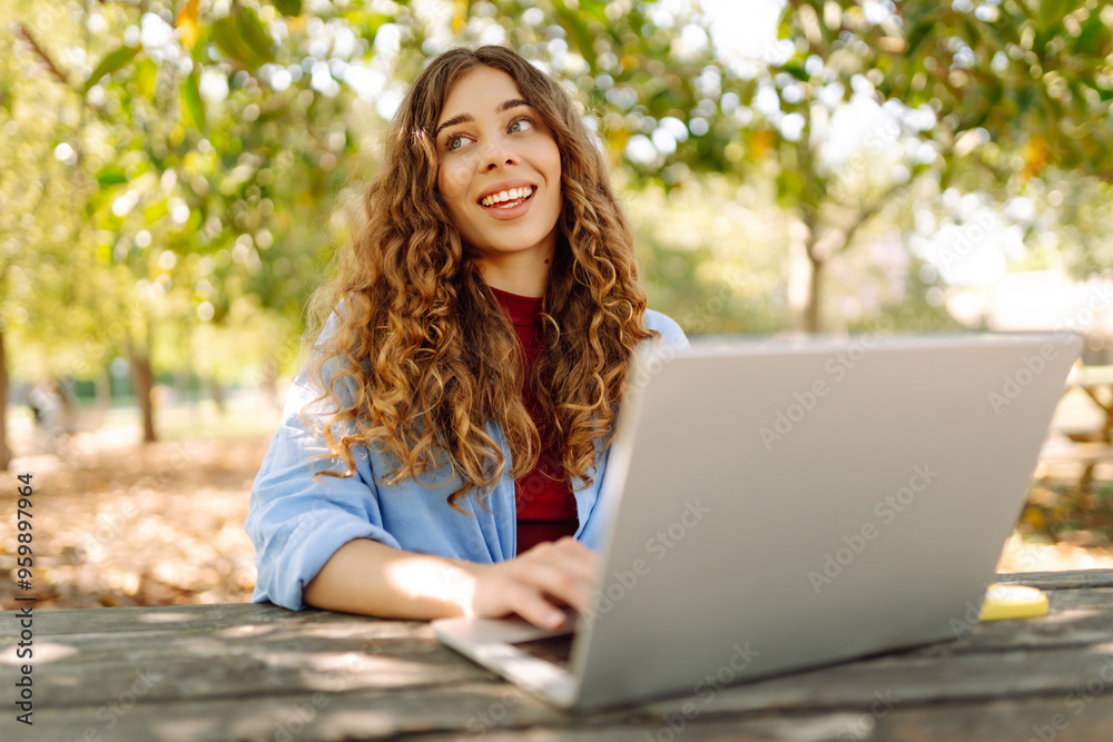 Young  freelancer woman in casual attire focused on laptop while sitting at a park table on a sunny day, enjoying a moment of productivity. E-learning concept. Remote work.