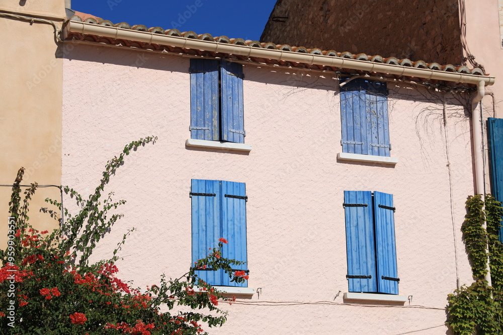 Closeup of colouful housefront adorned with bougainvillea flowers in bloom and featuring blue wooden window shutters in Mediterranean seaside town, southern France Closeup of colouful housefront adorned with bougainvillea flowers in bloom and featuring blue wooden window shutters in Mediterranean seaside town, southern France