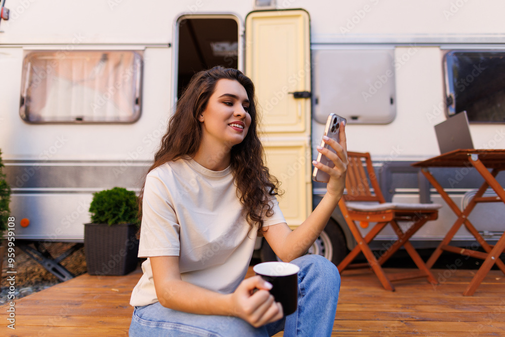 Young pretty woman use phone while have morning coffee sitting near camper Young pretty woman use phone while have morning coffee sitting near camper