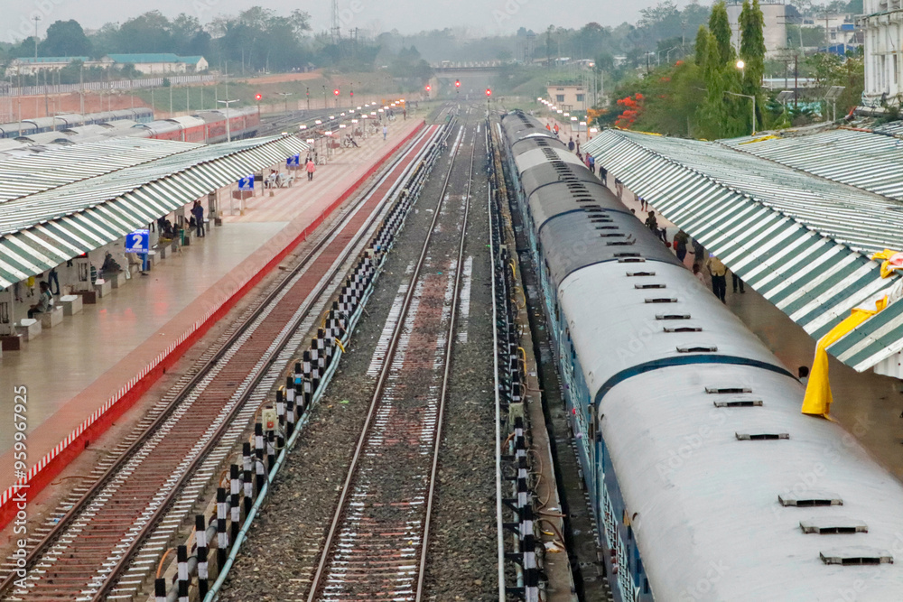 Fototapeta premium Railway station with multiple tracks, platforms, and stationary trains. Covered shelters over platforms, scattered passengers walking. Overcast sky, urban setting, showcasing transportation hub.