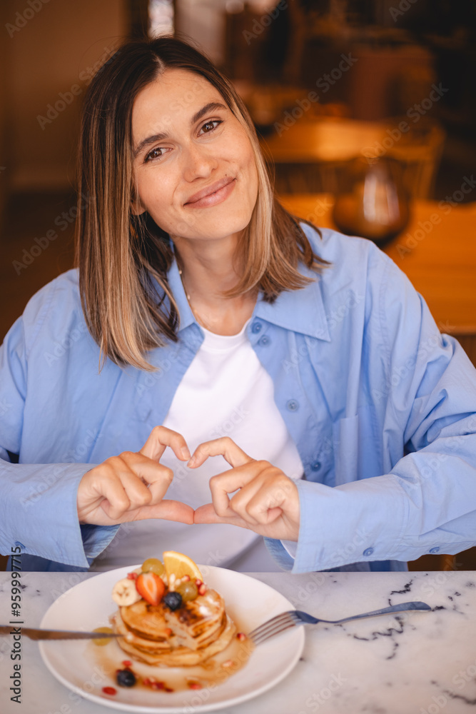 Laughing woman resting in cafe while enjoying tasty pancakes with fruit, maple syrup and show love heart gesture. Blonde short hair girl wear blue shirt and white t-shirt. Laughing woman resting in cafe while enjoying tasty pancakes with fruit, maple syrup and show love heart gesture. Blonde short hair girl wear blue shirt and white t-shirt.