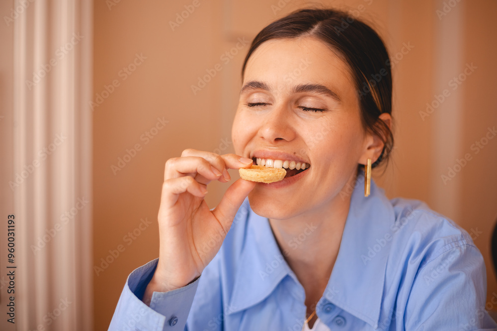 Happy woman eating cookie sitting in a restaurant. Positive woman bites cookie enjoys eating sweet food breaks diet. Woman wear blue shirt, low ponytail rest in cafe. Girl bite cake and look happy.