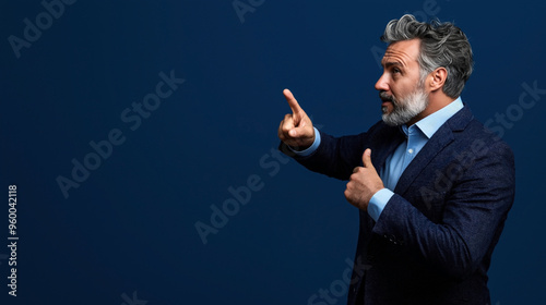 Mature man with gray hair and beard wearing a dark blue blazer and light blue shirt, pointing with index finger and thumb up against a solid dark blue background.