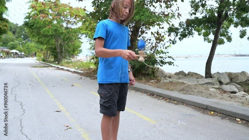 A young boy in a blue shirt plays with a kendama toy by the seaside, surrounded by trees and rocks. The calm water and peaceful setting enhance the relaxed outdoor activity