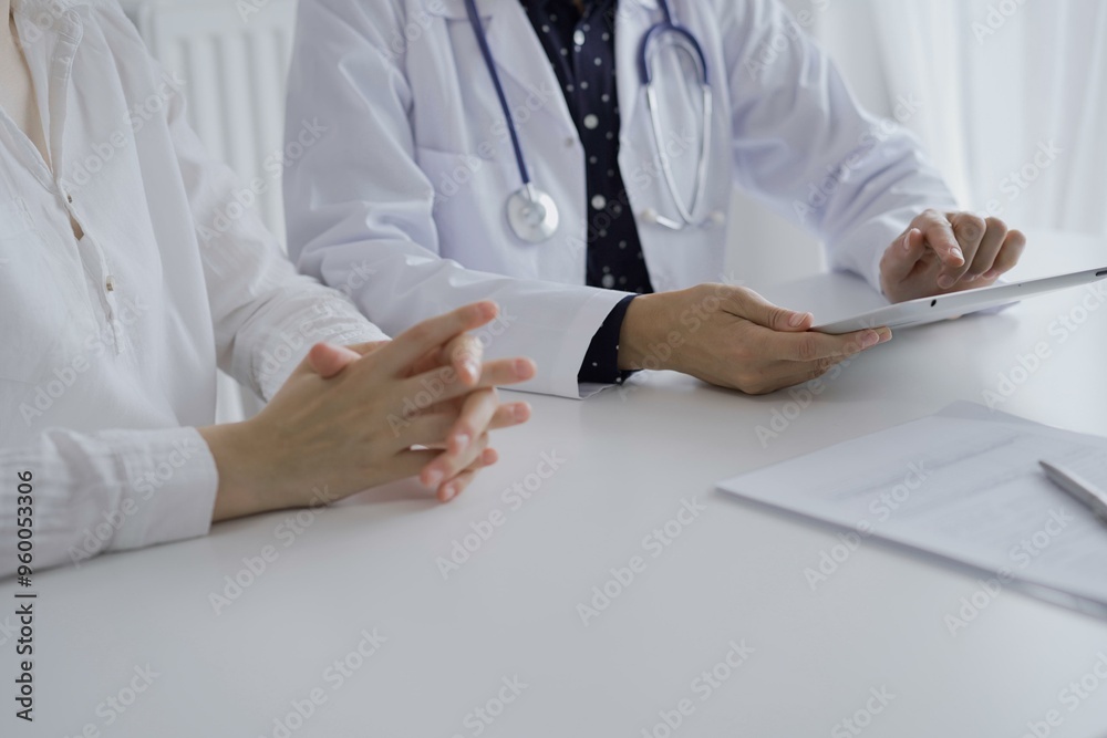 Doctor and a patient. The female physician, wearing a white medical coat over a dark blue dotted blouse, using tablet computer during a consultation in the clinic. Medicine concept
