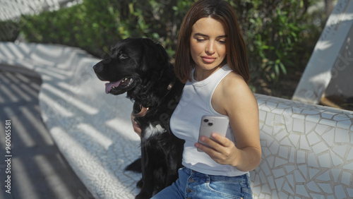 A young hispanic woman sitting on an outdoor bench in the city park, using her phone while her black labrador dog sits beside her.