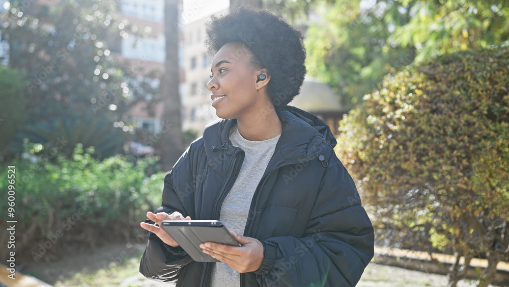 African american woman with curly hair enjoys music through wireless earbuds while holding a tablet outdoors. African american woman with curly hair enjoys music through wireless earbuds while holding a tablet outdoors.