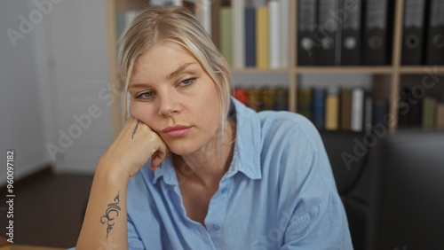 Young woman with blonde hair looking pensive in an office environment with books and folders in the background