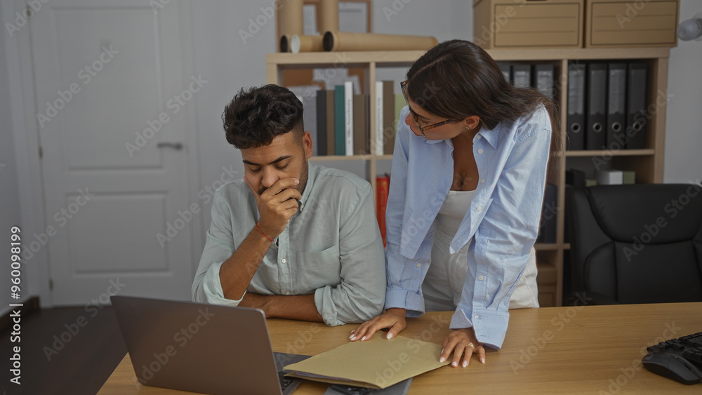 Woman discussing business documents with man in modern office workspace with organized shelves and laptop Woman discussing business documents with man in modern office workspace with organized shelves and laptop
