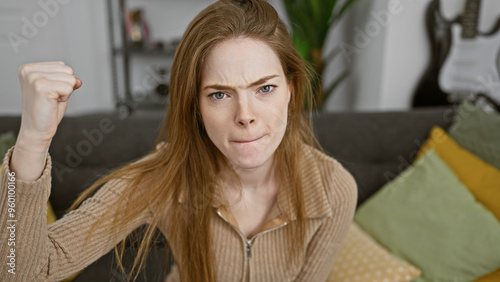 A frustrated young woman clenches her fist in a cozy living room, expressing annoyance or determination.