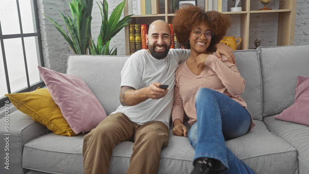 Interracial couple relaxing together on a grey couch in a cozy urban living room holding a remote Interracial couple relaxing together on a grey couch in a cozy urban living room holding a remote