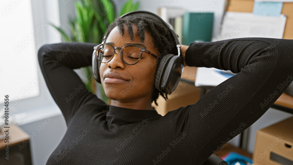 Fototapeta premium A relaxed african american woman with dreadlocks enjoys music on headphones in a modern office setting.