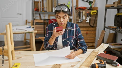 South asian woman checks phone while working in a carpentry studio with protective goggles.