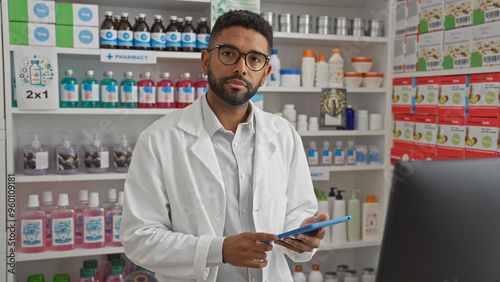 African american pharmacist with beard stands in pharmacy holding a tablet, surrounded by shelves of medicine and health products.