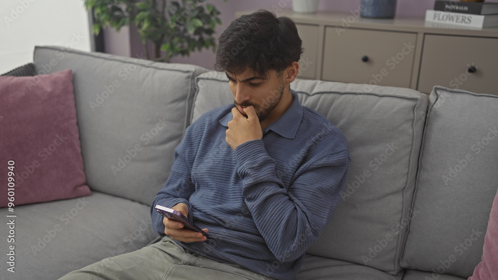 Thoughtful young man with beard using smartphone on couch in modern living room interior. Thoughtful young man with beard using smartphone on couch in modern living room interior.