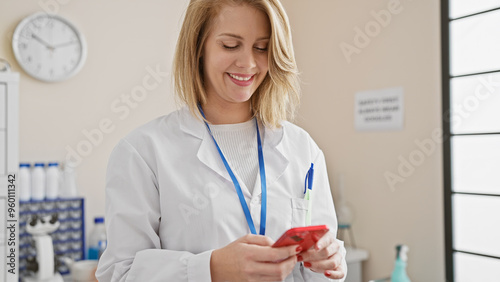 Smiling blonde woman in lab coat using smartphone in clinic