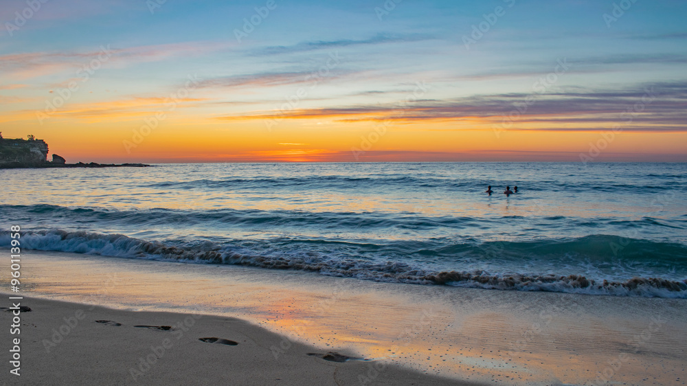 Sunrise view on the beach in Sydney, Australia. In the background, the sky with the sun rising with orange and blue colors, while it contrasts with the sea and the silhouettes of people bathing. Sunrise view on the beach in Sydney, Australia. In the background, the sky with the sun rising with orange and blue colors, while it contrasts with the sea and the silhouettes of people bathing.